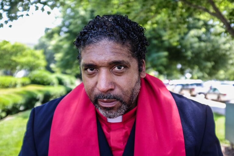 Rev. William J. Barber II standing outside wearing the black suit of a pastor and a red scarf. Trees in the background.