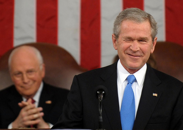 George W. Bush in a suit and blue tie winking in front of a large American flag while Dick Cheney sits behind him applauding.