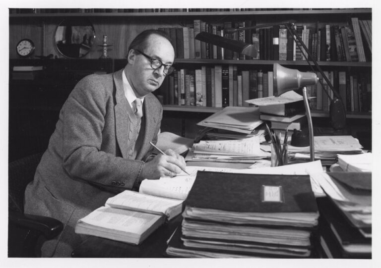 Black-and-white photograph of novelist Vladimir Nabokov sitting at a desk covered with books and papers, writing in a notebook.