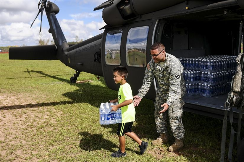 Uniformed military member hands child a pallet of bottled water next to a helicopter.