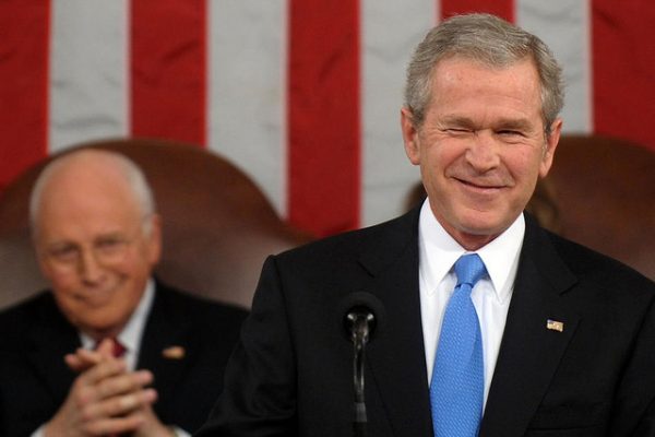 George W. Bush in a suit and blue tie winking in front of a large American flag while Dick Cheney sits behind him applauding.